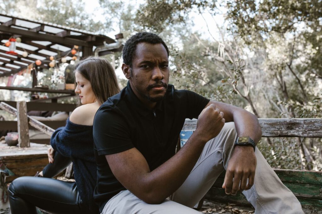 A thoughtful man sits outdoors, with a woman sitting distantly behind him.