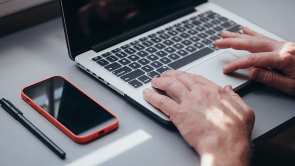 Hands typing on a laptop with a smartphone and pen on the side, perfect for tech and work themes.